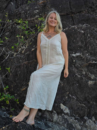 A woman wearing a sleeveless, off-white maxi dress with a lace-up front, posing on dark volcanic rocks outdoors.