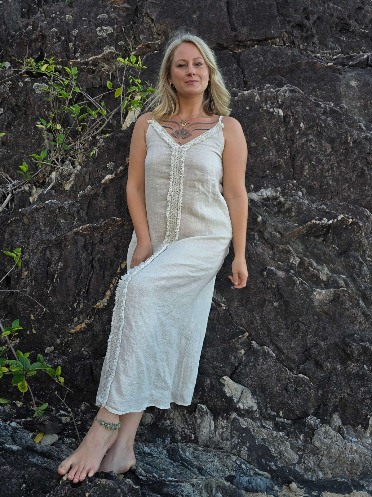 A woman with blonde hair stands barefoot against a dark rocky cliff, wearing a long sleeveless cream linen dress with a buttoned front and gathered waist.