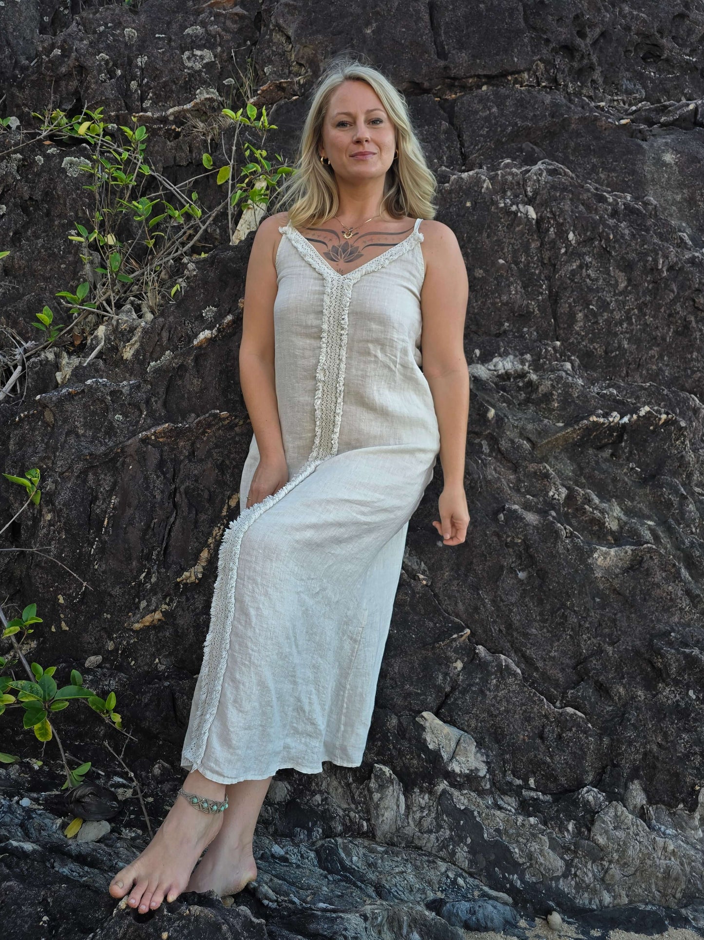 A woman with blonde hair stands barefoot against a dark rocky cliff, wearing a long sleeveless cream linen dress with a buttoned front and gathered waist.