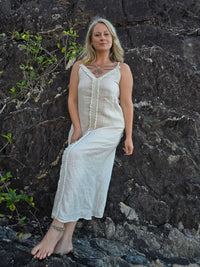 A woman with blonde hair stands barefoot against a dark rocky cliff, wearing a long sleeveless cream linen dress with a buttoned front and gathered waist.