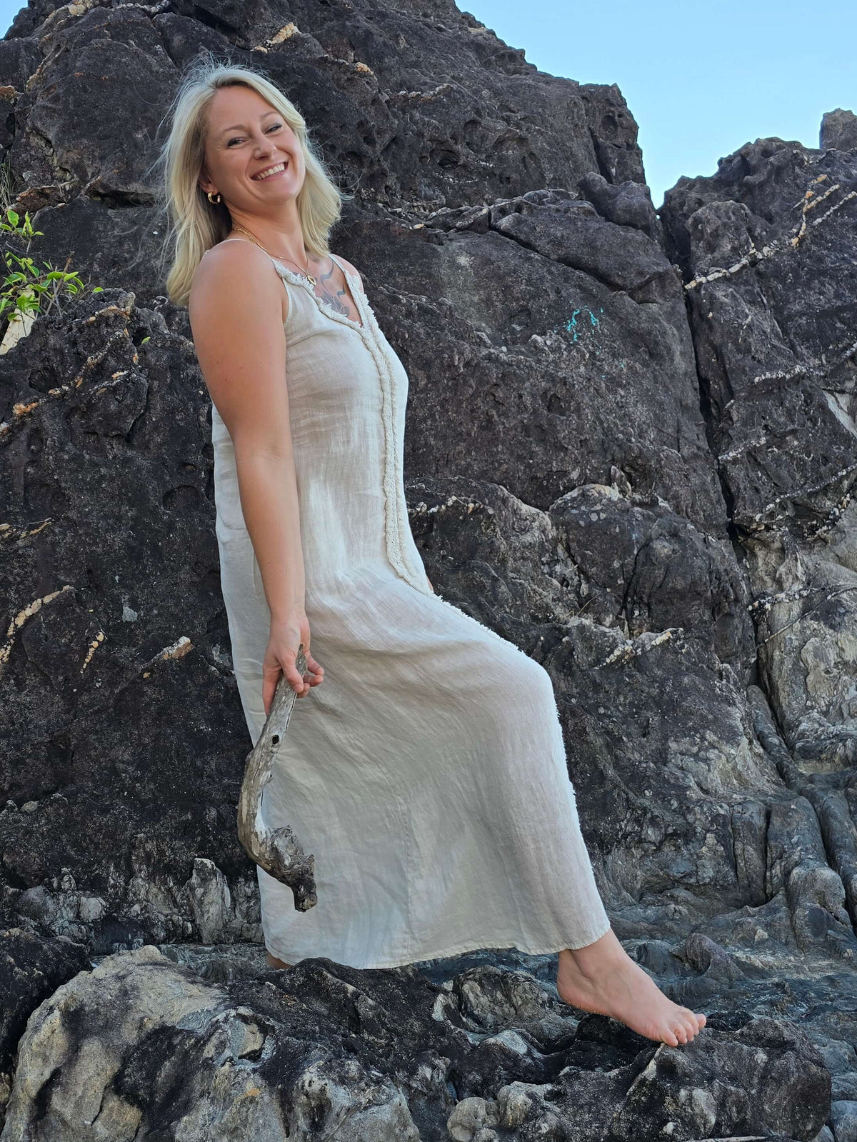 A woman wearing a light beige linen maxi dress sits on dark volcanic rocks against a blue sky background.