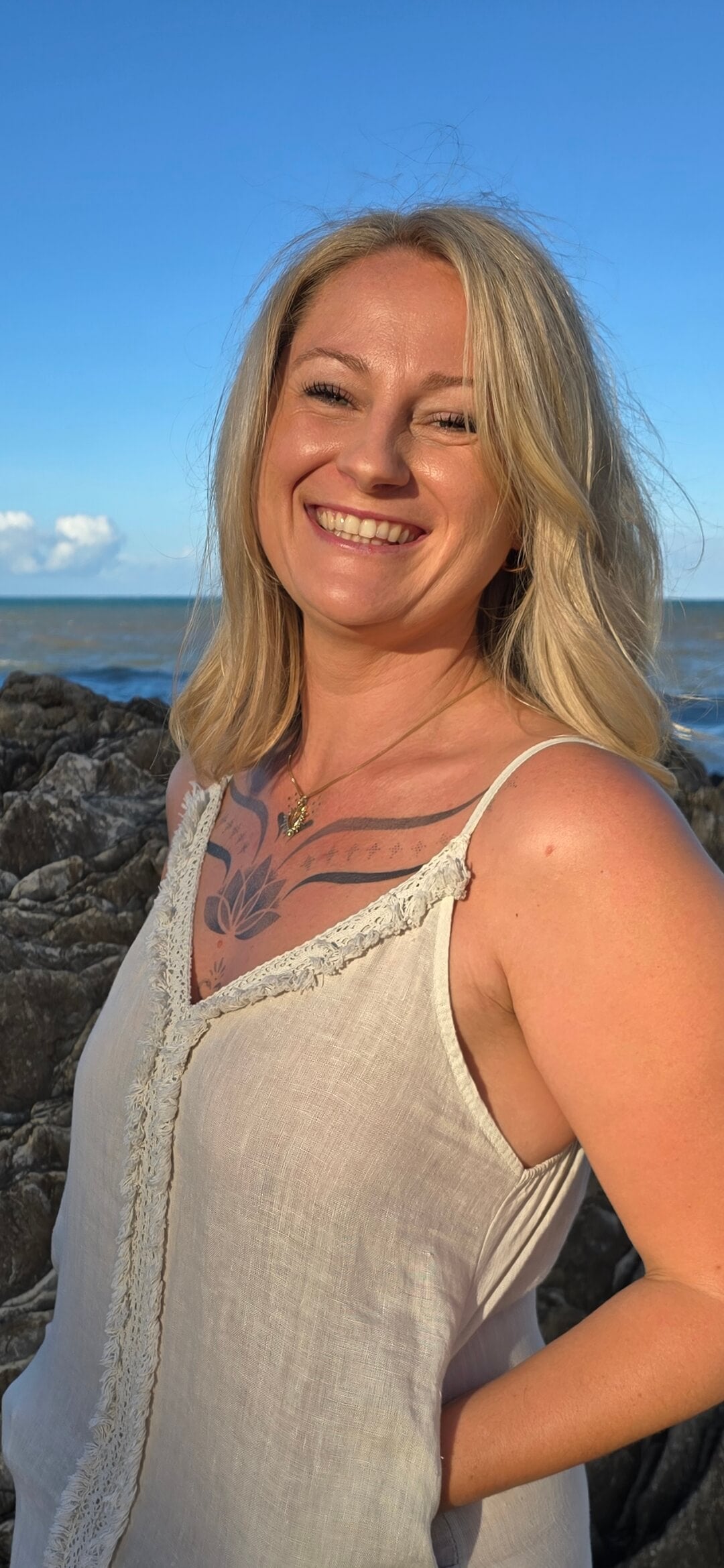 A smiling blonde woman wearing a light beige sleeveless top stands outdoors by the ocean with rocks and blue sky in the background.