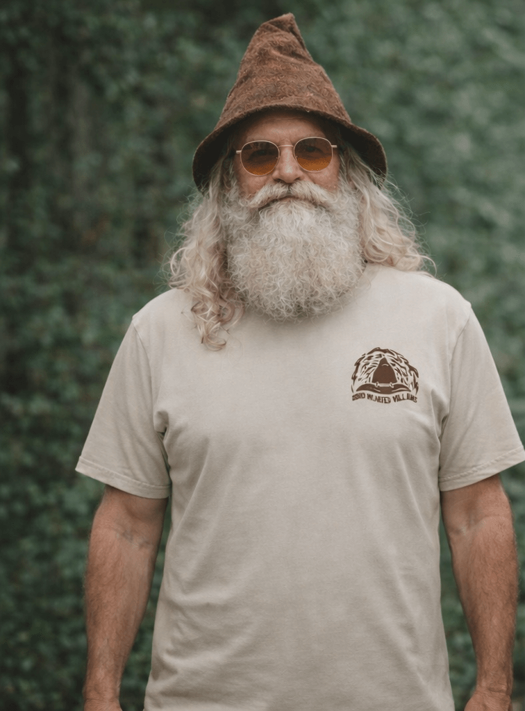 Man wearing a beige t-shirt with a gnome graphic and text, standing against a green leafy background.