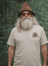 Man wearing a beige t-shirt with a gnome graphic and text, standing against a green leafy background.