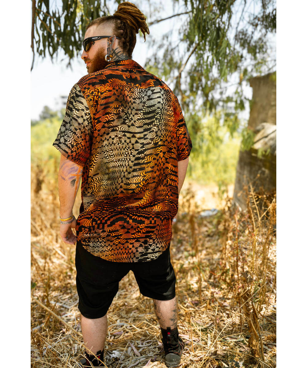 Man wearing a patterned shirt and sunglasses standing in a natural setting with trees and dry grass.