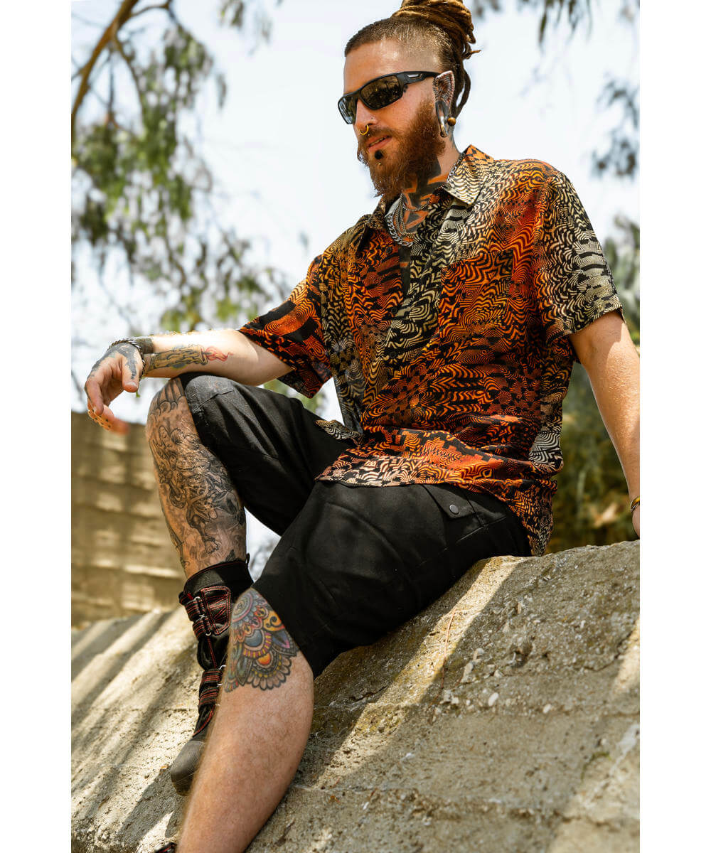 Man with tattoos wearing a patterned shirt and sunglasses, sitting outdoors.
