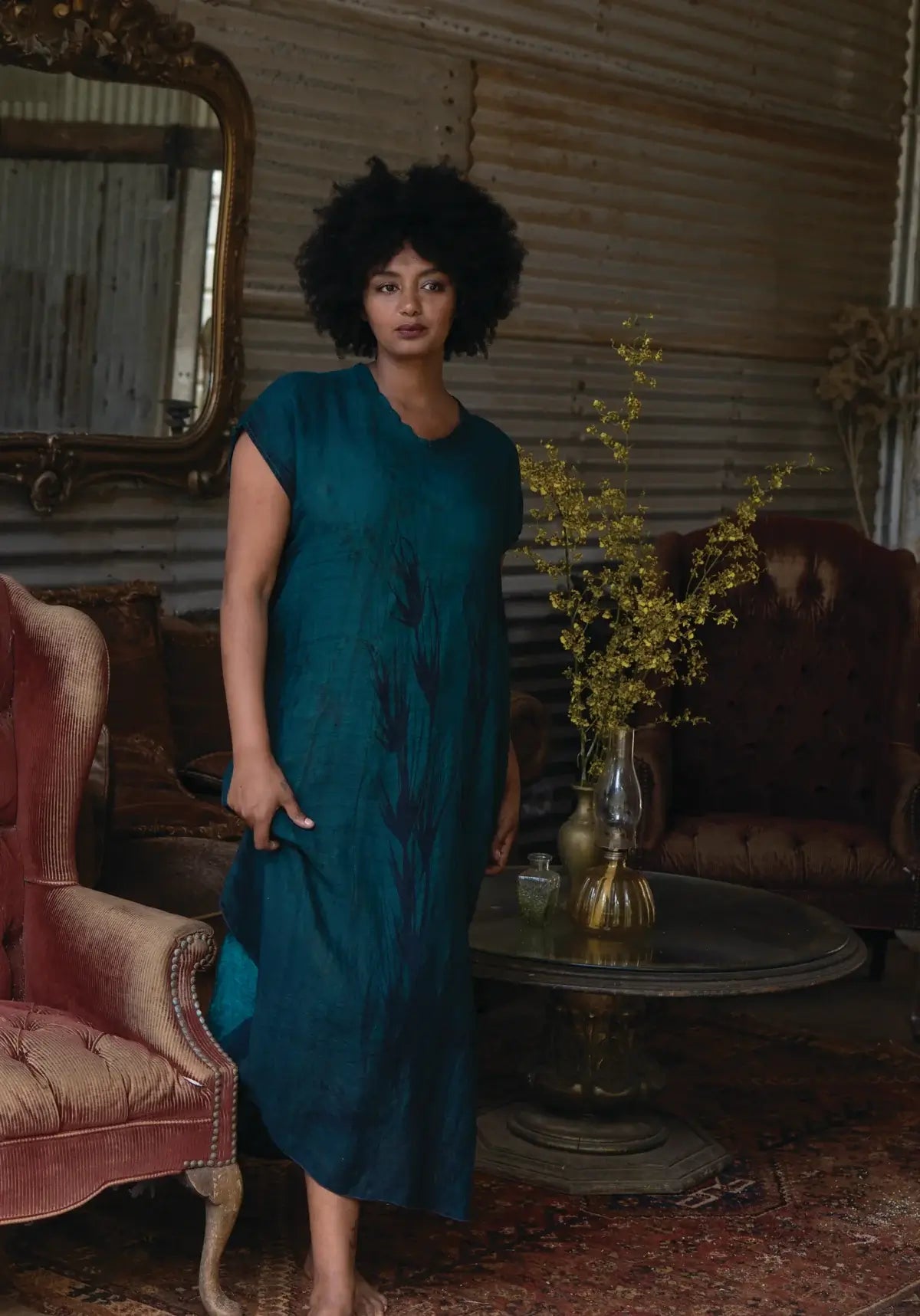 Woman in a teal dress standing in a vintage-style room with a mirror and floral arrangement.
