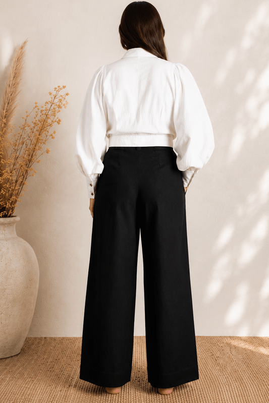 Person wearing a white blouse and black pants standing in a room with a vase and plant.