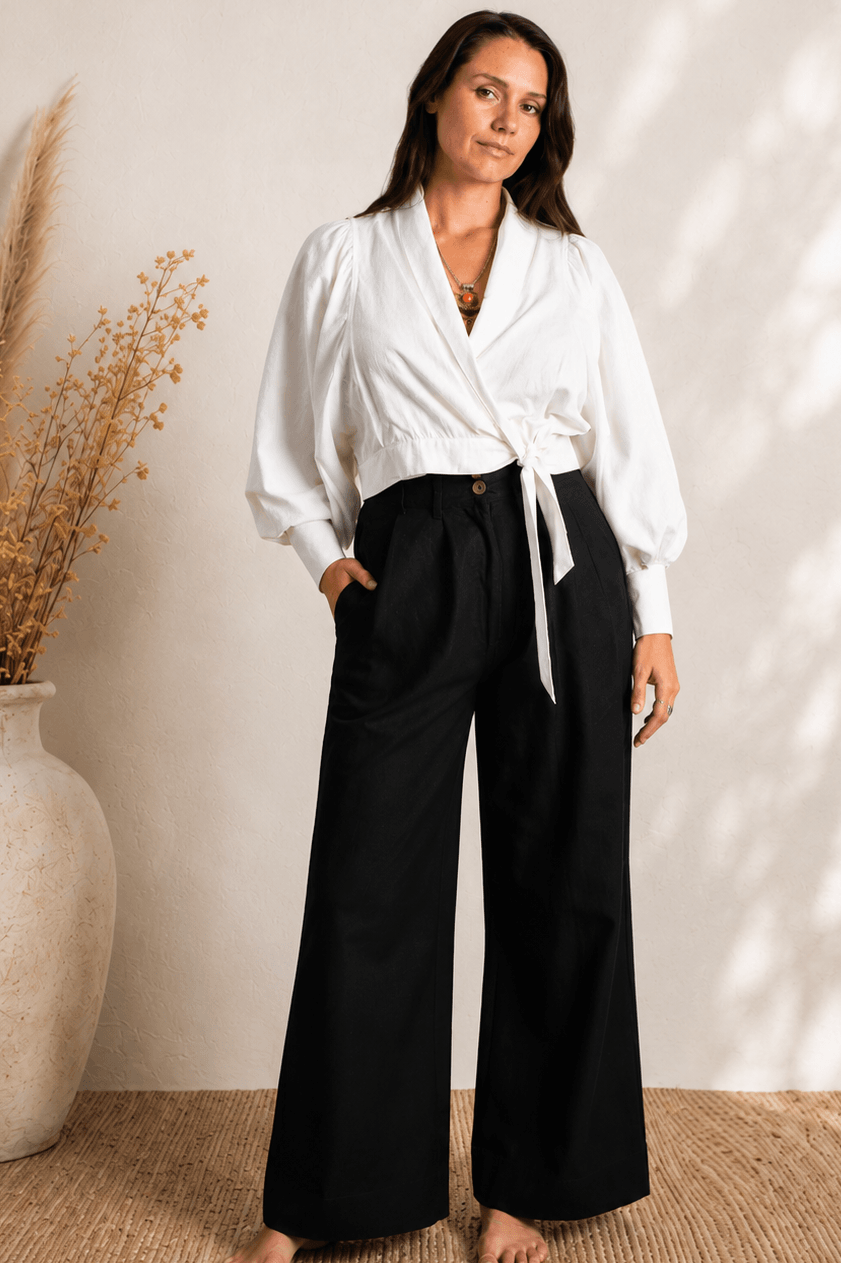 Woman wearing a white blouse and black pants standing against a neutral wall with a vase of dried plants.