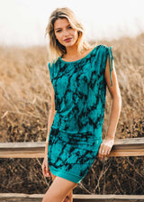 Woman wearing a teal dress with black patterns standing outdoors against a natural background.