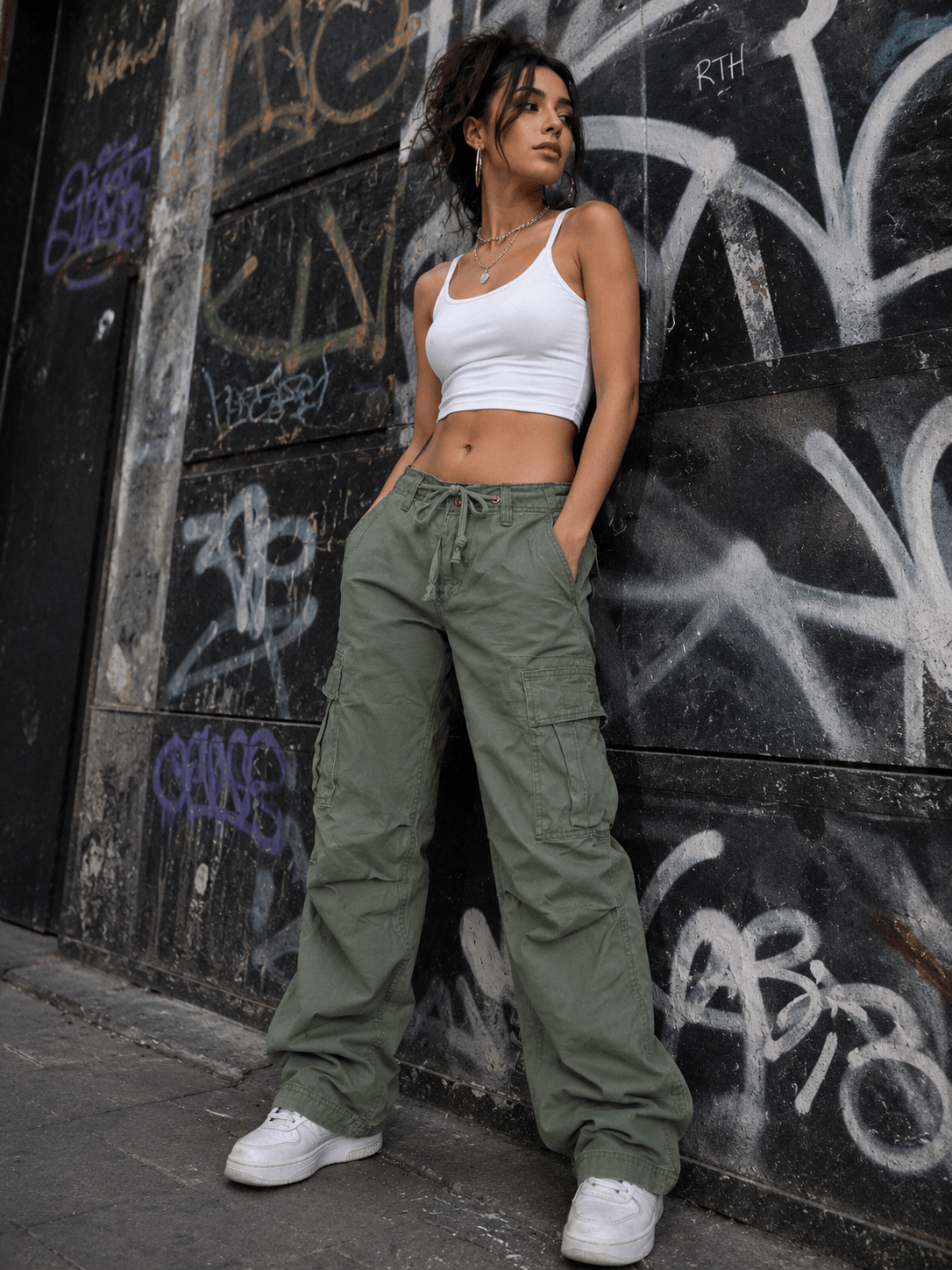 Woman in white crop top and green cargo pants leaning against a graffiti-covered wall.