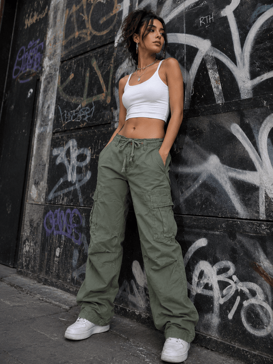 Woman in white crop top and green cargo pants leaning against a graffiti-covered wall.