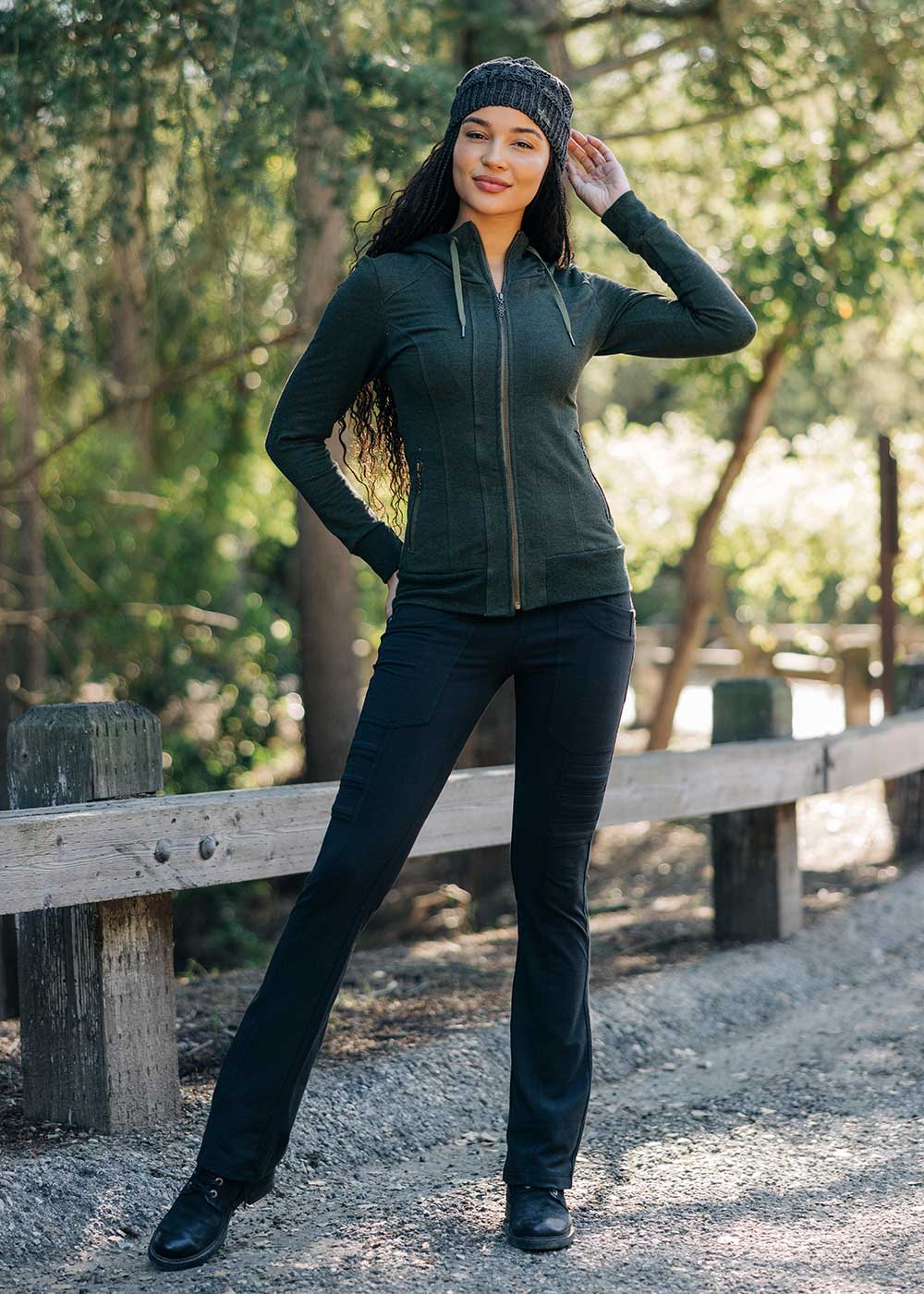 Woman wearing a green jacket and black pants standing outdoors near a wooden fence.