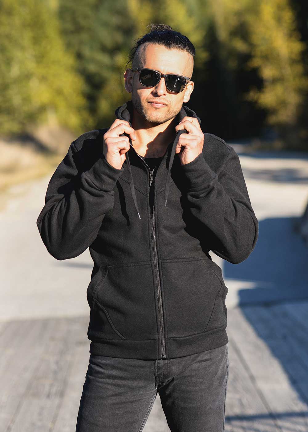 Man wearing a black hoodie and sunglasses outdoors with trees in the background