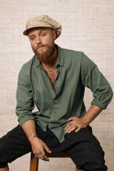 Man wearing a green shirt and beige cap sitting on a stool against a textured wall.
