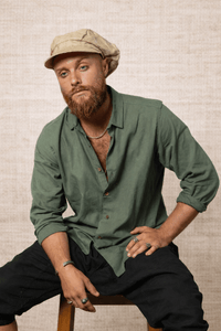 Man wearing a green shirt and beige cap sitting on a stool against a textured wall.