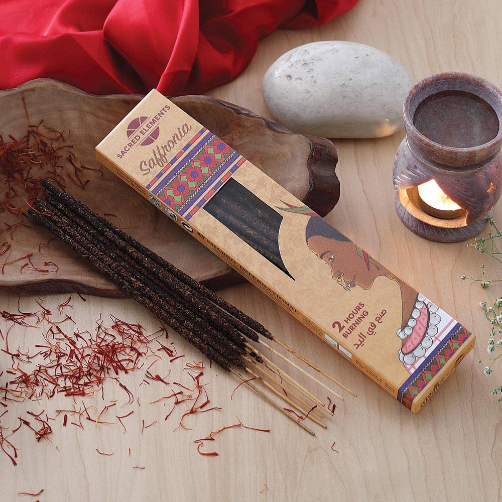 A box of Sacred Heart Salvia incense sticks lies on a light wood surface beside several dark incense sticks, a wooden tray, scattered red incense shreds, a smooth stone, and a small lit candle holder.