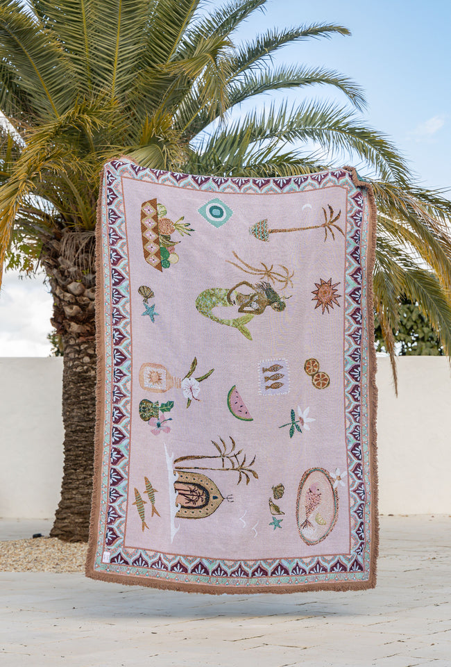 Decorative towel with mermaid and shell patterns hanging against a palm tree and white wall background.