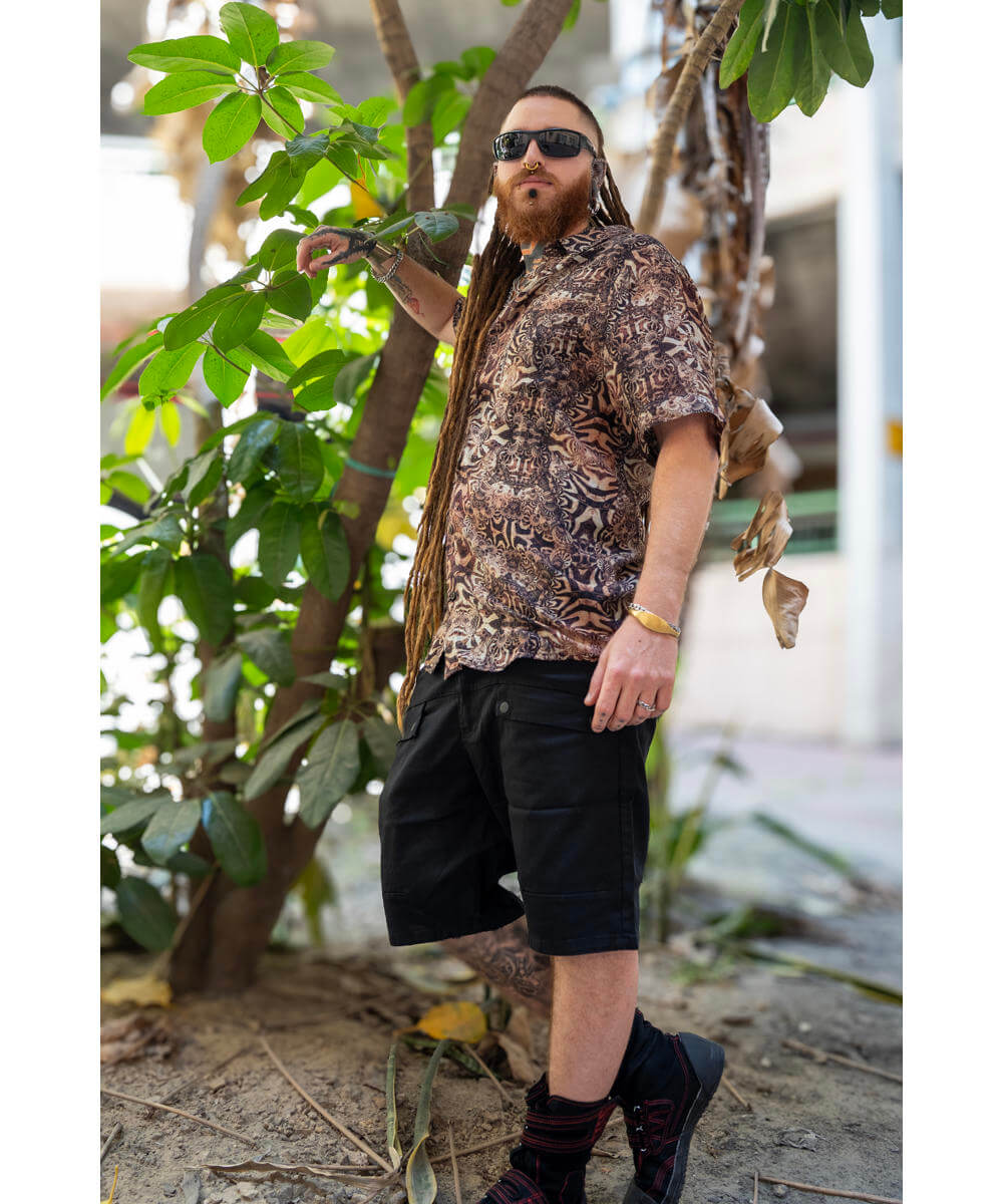 Man in a patterned shirt and black shorts standing among green foliage.