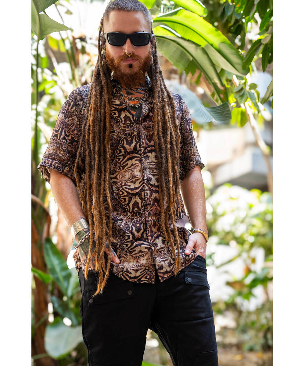 Man with dreadlocks wearing a patterned shirt and sunglasses in a tropical setting